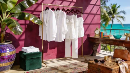 A sleek brass clothing rack holds white resort wear garments, including a blazer, blouse, and jumpsuit, against a magenta wall next to an open terrace with an ocean view.