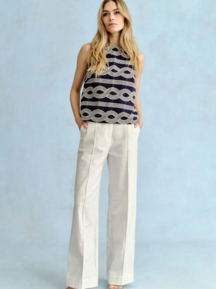 A woman stands against a light blue background wearing soft white Cartolina Beverly wide-leg trousers paired with a sleeveless navy blue top featuring a white wavy embroidered chain link pattern.