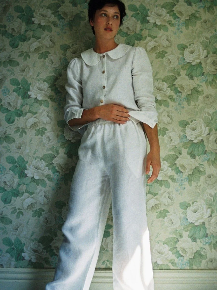 Model posing against a floral wall wearing the white Má + Lin Belgian linen trousers and long-sleeve blouse.