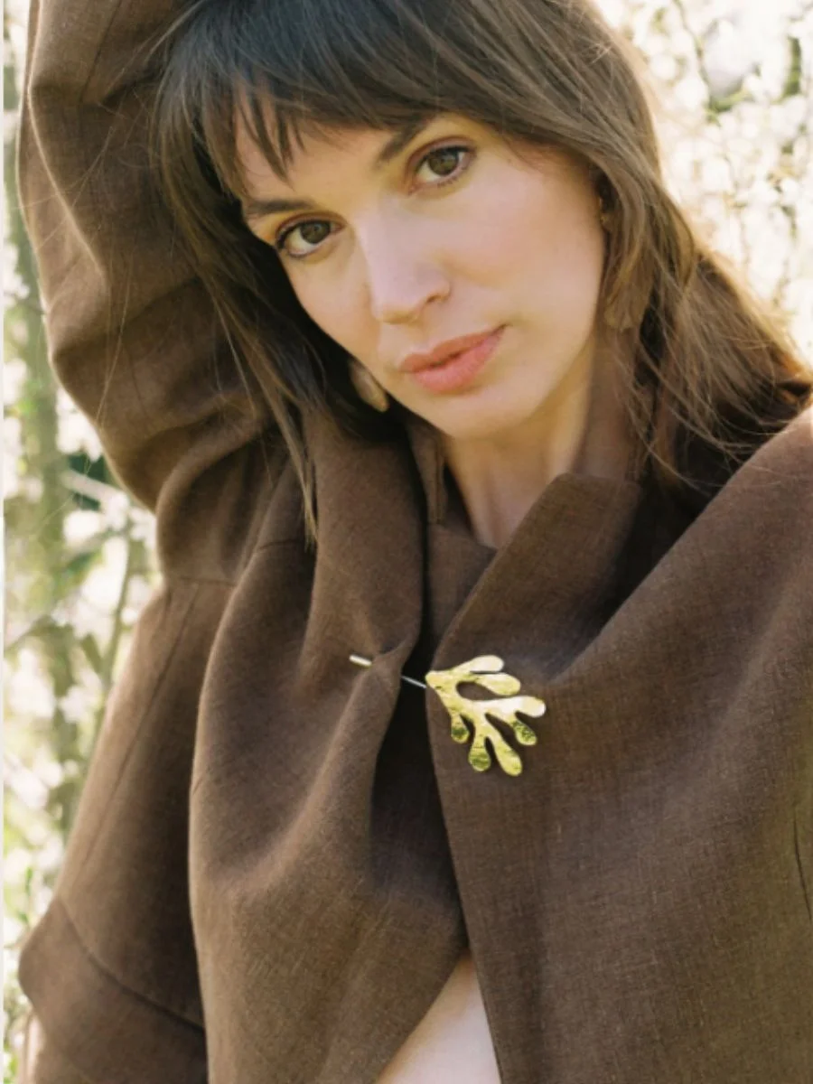 Close-up of the cinnamon Má + Lin blazer pinned shut at the upper lapel with an organic gold brooch.