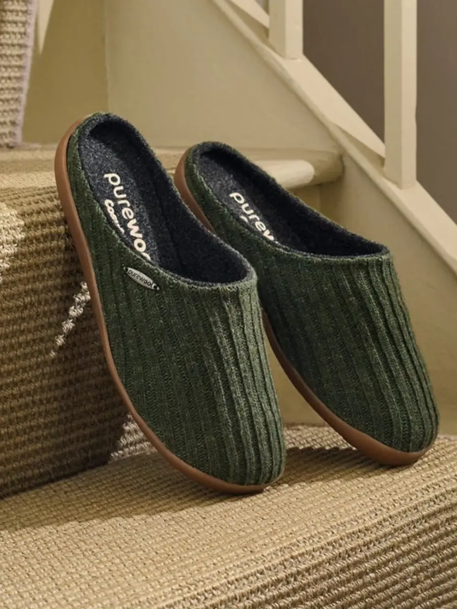 A pair of olive green ribbed wool mule slippers with a dark grey inner lining and brown rubber outsoles, resting on carpeted indoor stairs.