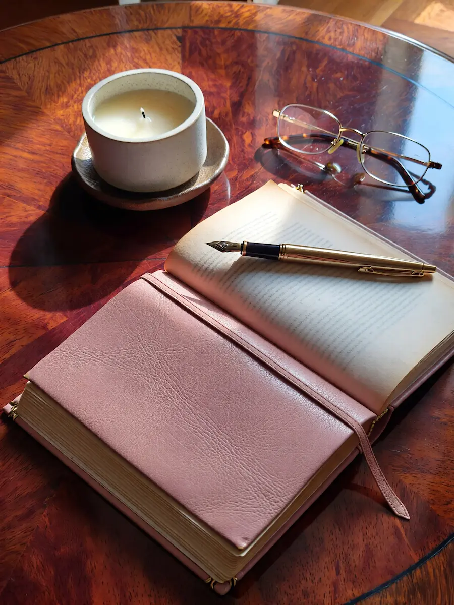 An open pink leather journal with a gold fountain pen resting on the pages next to a lit ceramic candle and reading glasses on a polished wood table.