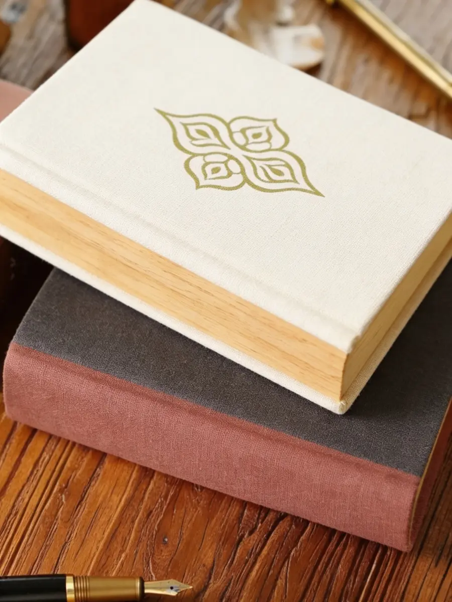 Two fabric-bound hardcover journals stacked on a rustic wooden desk next to a fountain pen.