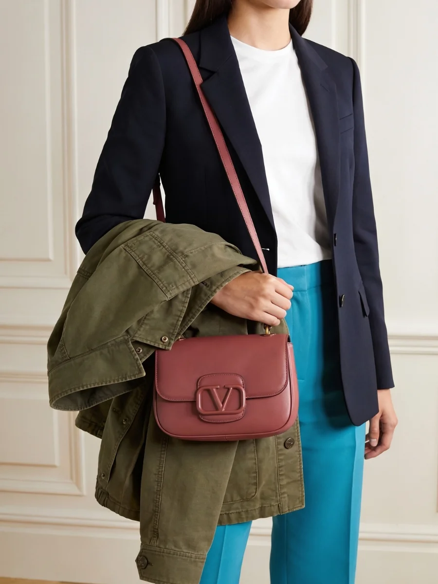 A woman styling a luxury burgundy leather shoulder bag with a dark blue blazer, an olive green utility jacket draped over her arm, and vibrant teal trousers.