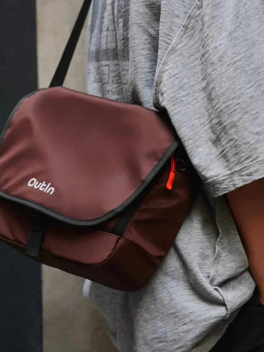A close-up shot of a person wearing an amber brown nylon messenger bag cross-body over a grey t-shirt, showing the white brand logo on the front flap and an orange zipper pull.
