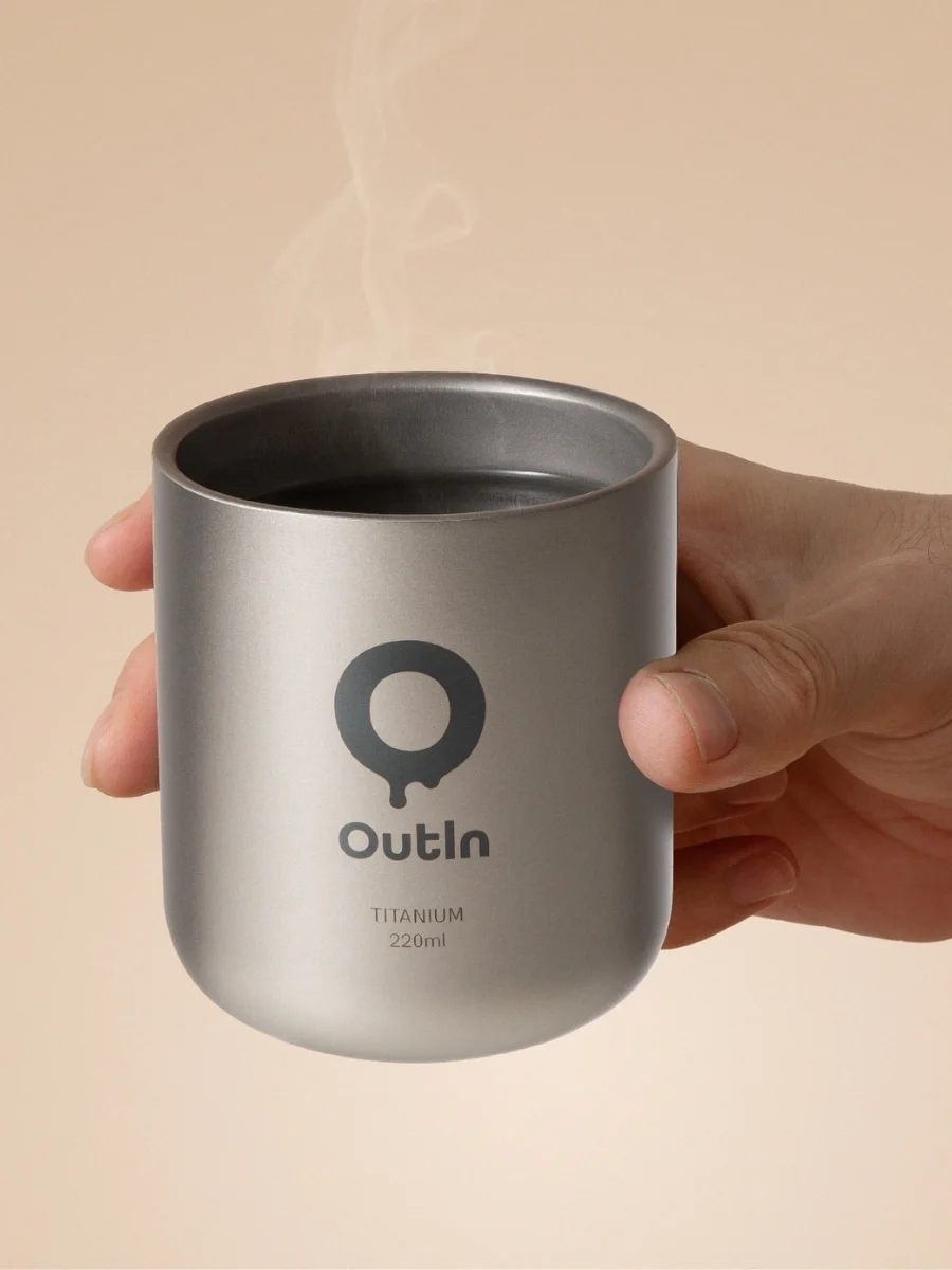 A hand holding a matte space grey double-wall titanium coffee cup with rising steam, showing the dark grey brand logo and the text TITANIUM 220ml on the side against a solid beige background.
