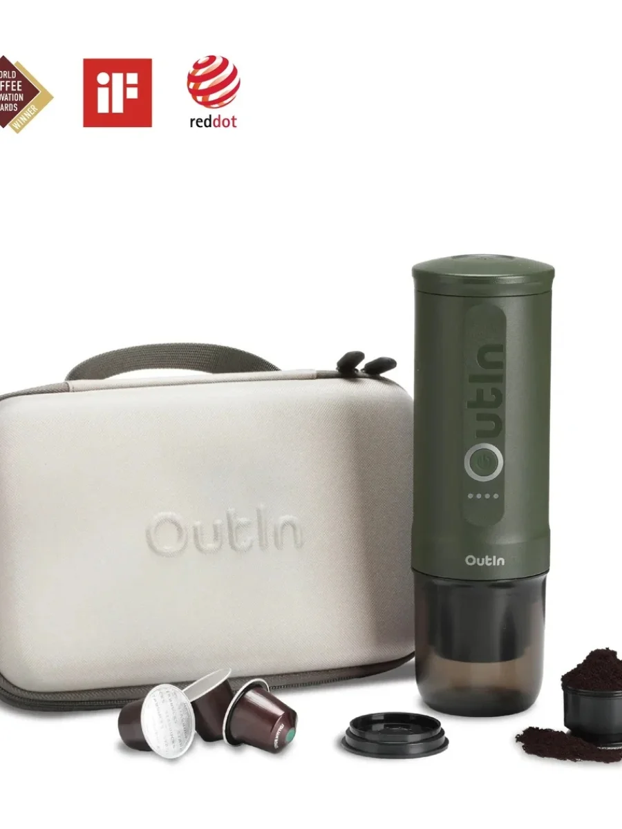 A forest green OutIn Nano portable electric espresso machine stands upright next to a matching hard-shell grey zip travel case embossed with the OutIn logo. In the foreground, three coffee capsules, a black cup lid, and a small measuring scoop filled with ground coffee are arranged on a clean white surface.