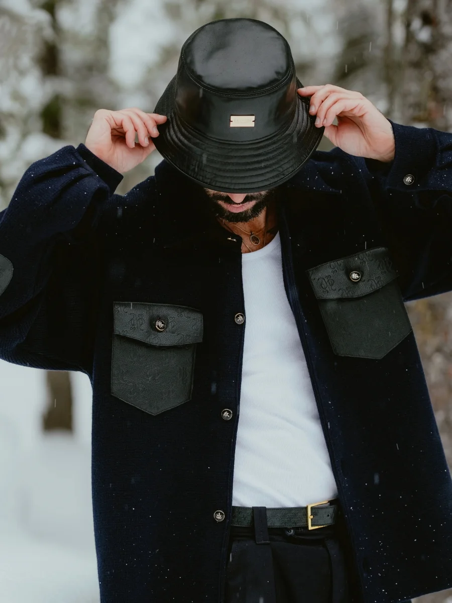 A man standing in a snowy environment adjusting a black leather bucket hat featuring a rectangular front plaque while wearing a heavy dark navy button-up shirt jacket over a white top.
