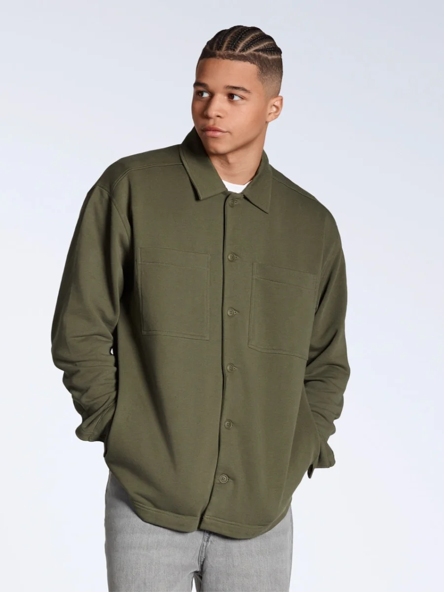 A man with a braided fade haircut stands against a plain white background, looking off-camera. He is wearing an olive green, buttoned-down organic cotton midweight overshirt over a white t-shirt, paired with light gray denim jeans.