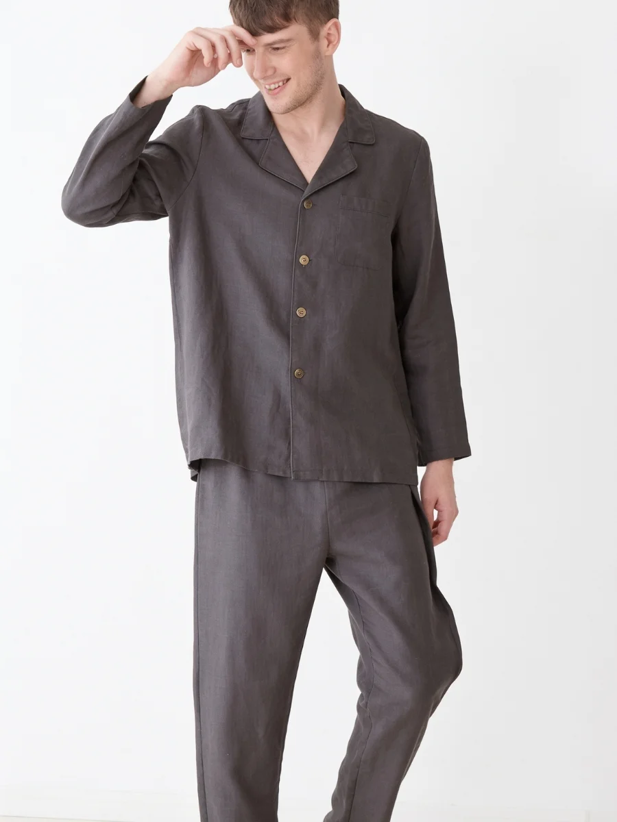 A man standing against a white wall wearing a dark grey long-sleeve linen pajama set featuring a button-down front and loose-fitting trousers.