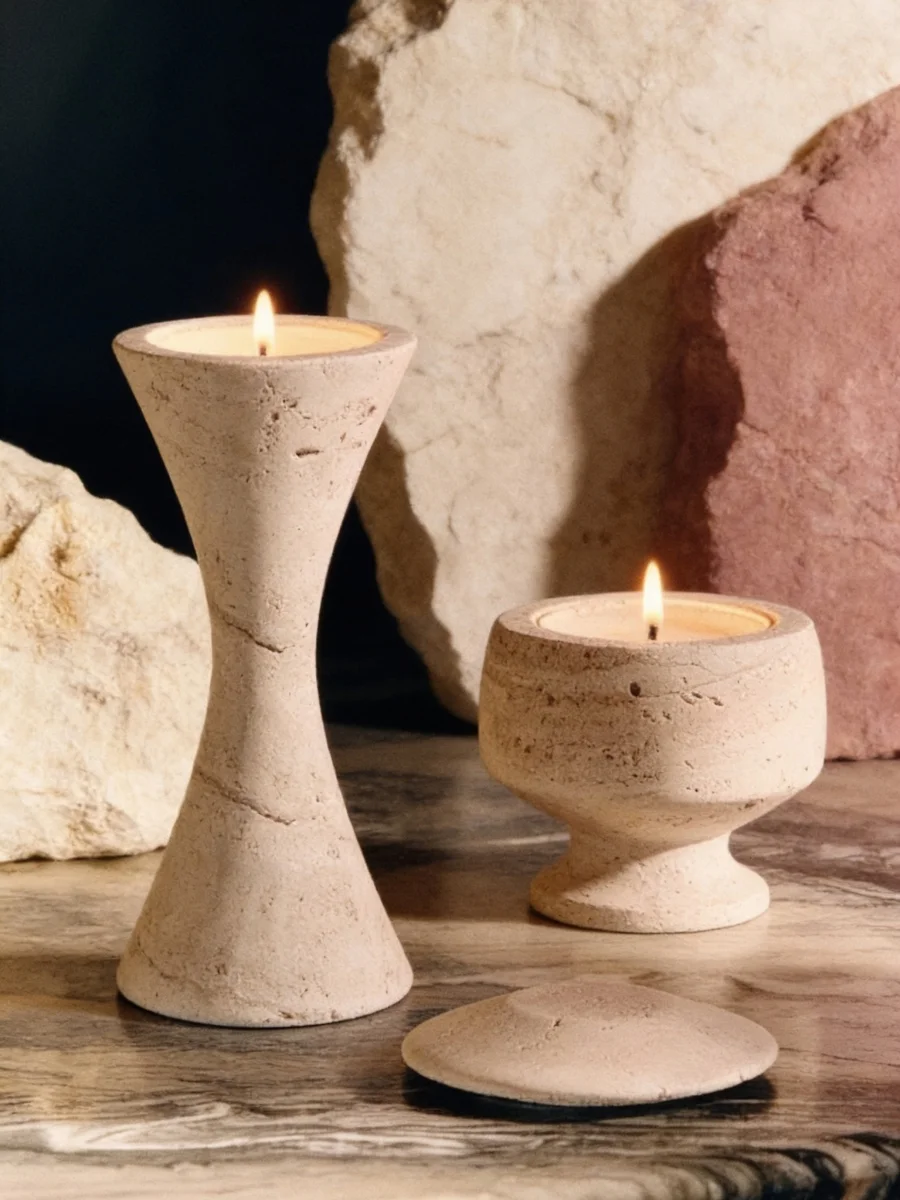Two unpolished travertine stone candle holders with lit wicks on a dark marble tabletop.