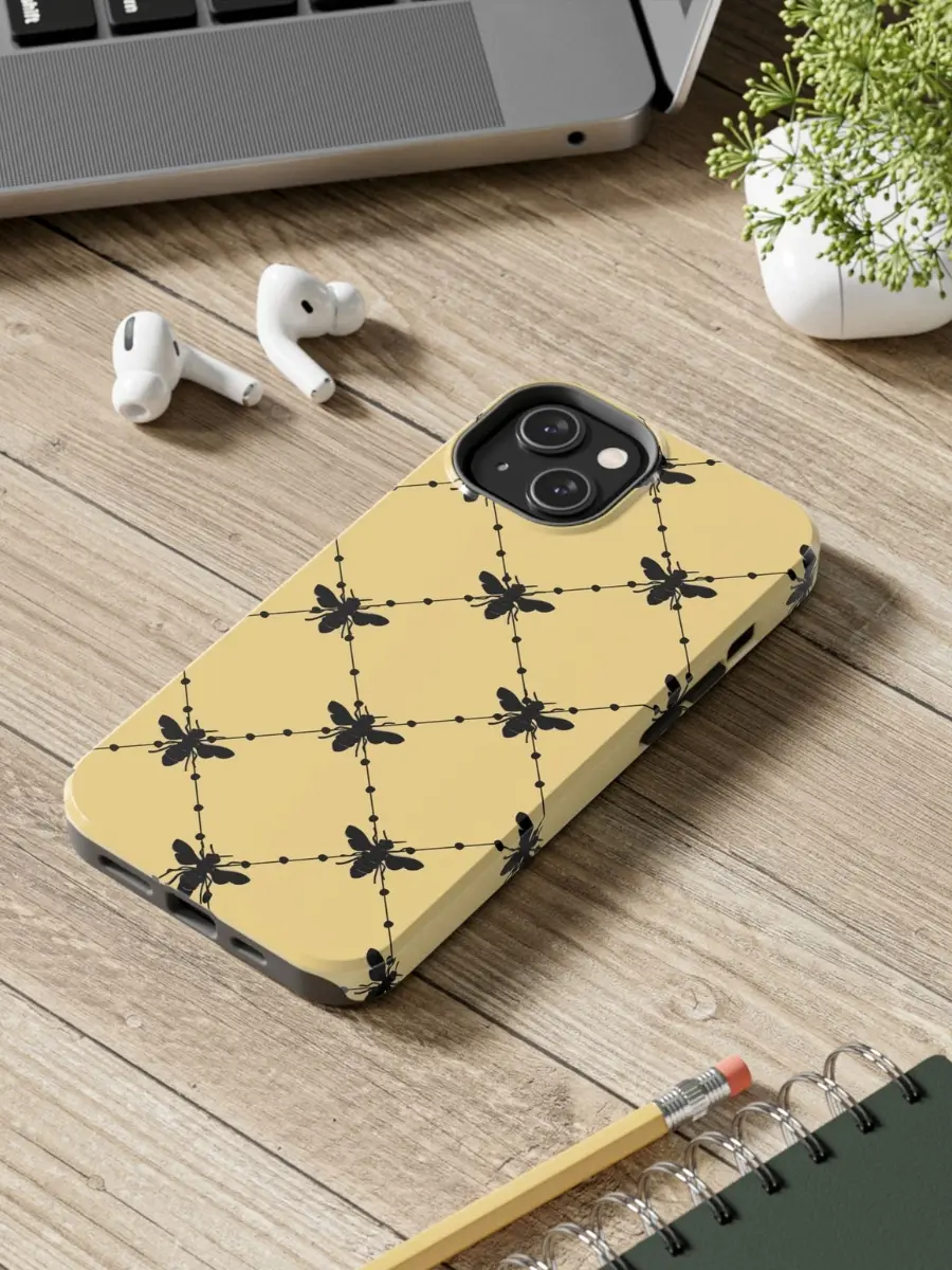 Beige phone case with a black bee and dotted diamond grid pattern on a wooden desk next to wireless earbuds and a laptop.