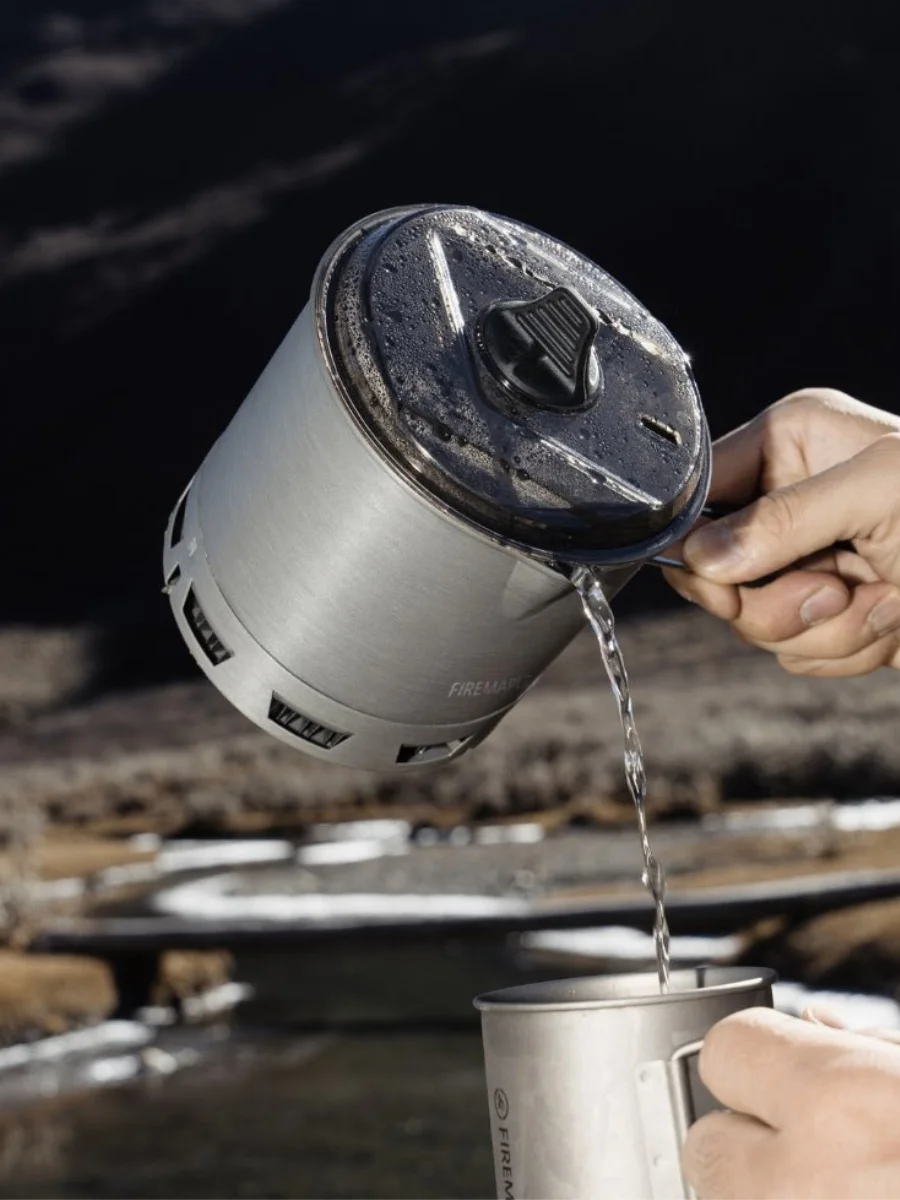 A close-up of a person pouring hot water from a grey camping pot into a metal mug in a sunny mountain landscape.