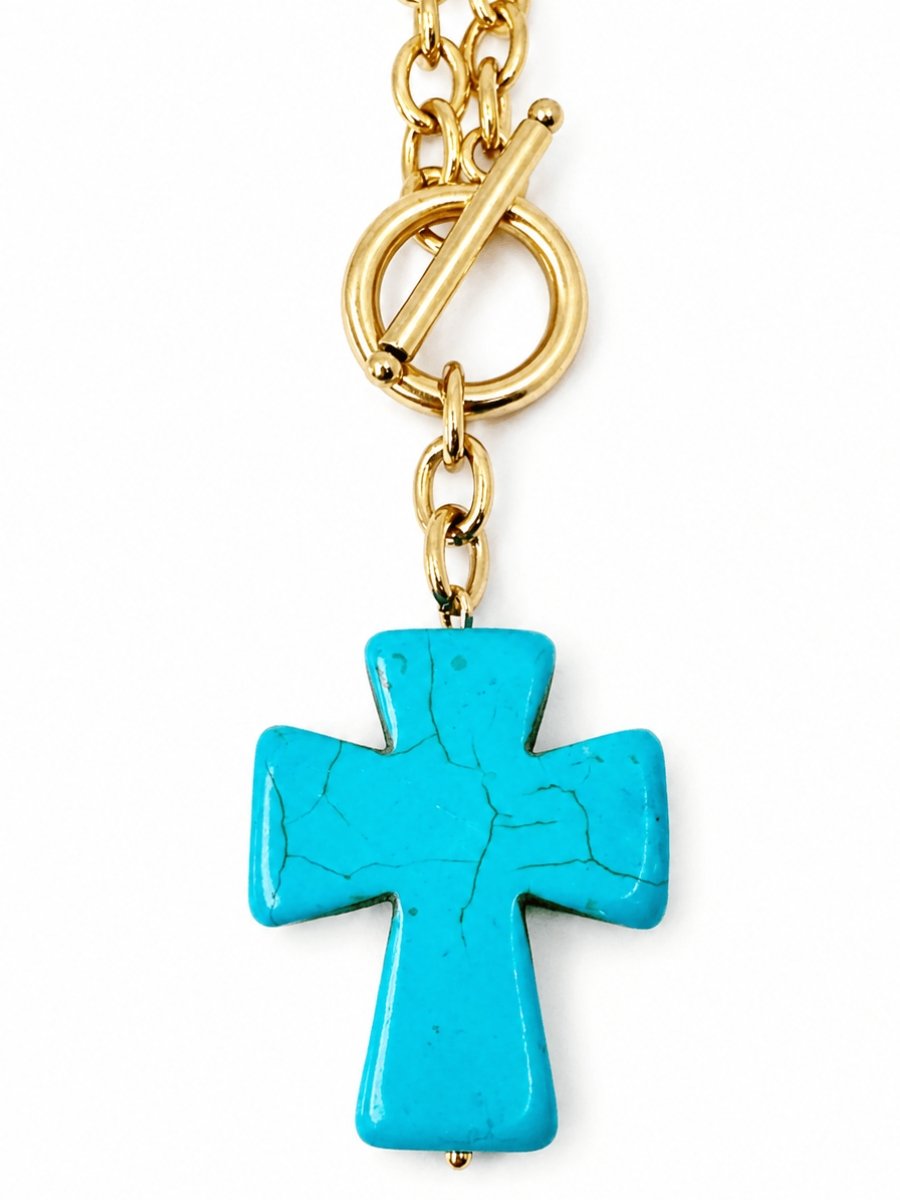 Gold plated chain necklace with a front toggle closure holding a turquoise blue howlite stone cross pendant on a plain white background.