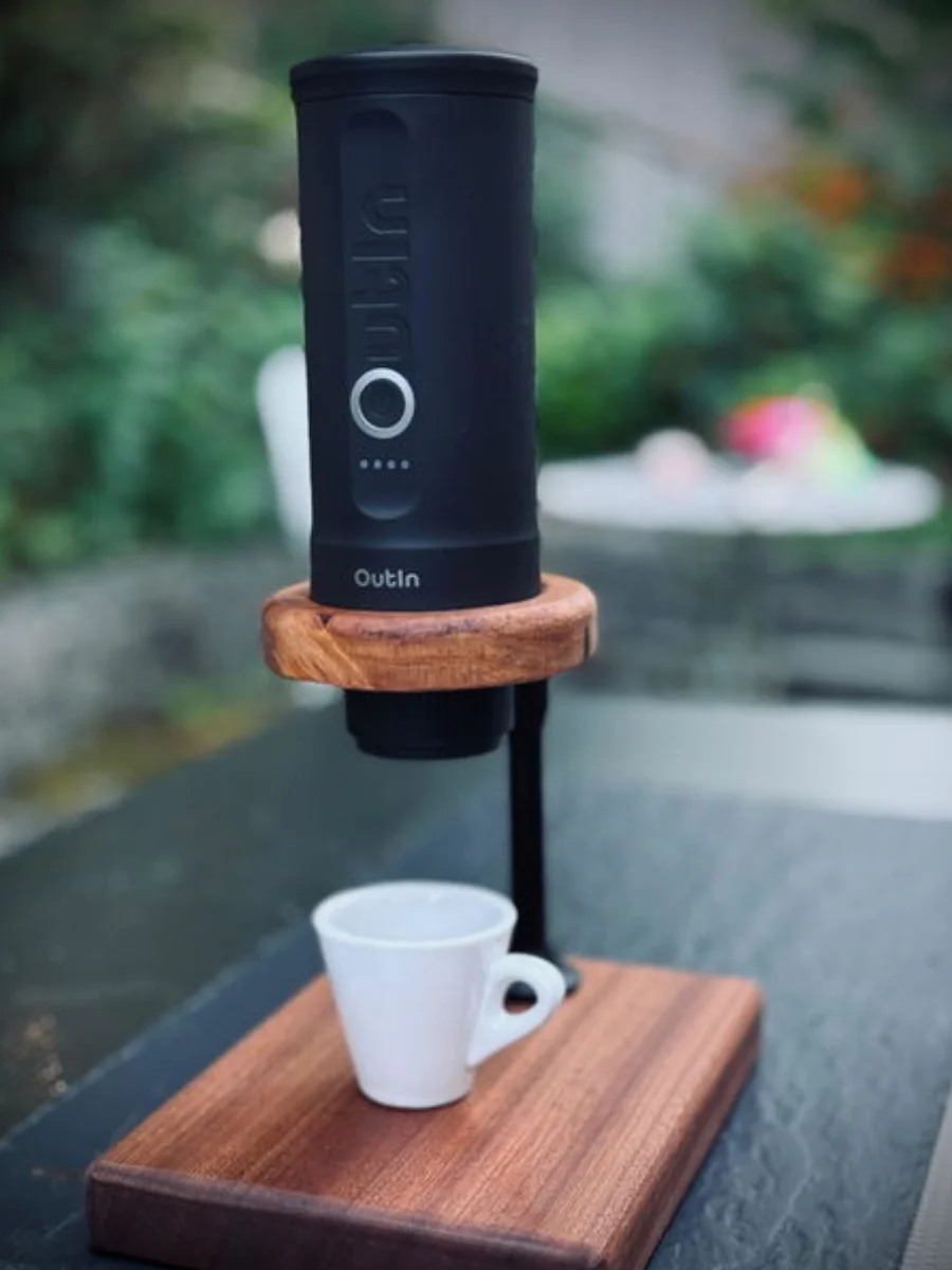 Portable espresso machine set up on a wooden counter stand with an empty white espresso cup underneath against a blurred green outdoor backdrop.
