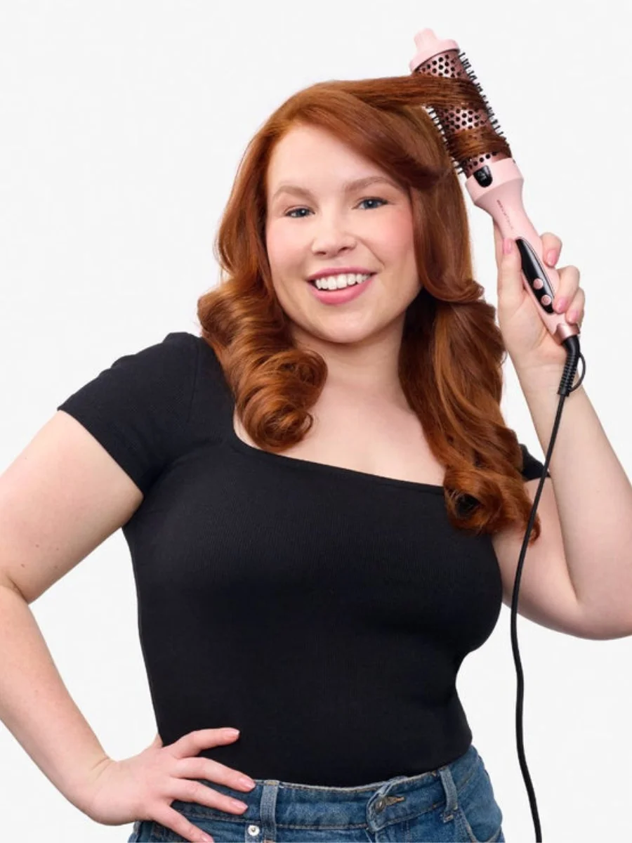 Smiling woman with long red wavy hair using a pink and black ionic thermal round brush to style her bangs on a plain white background.
