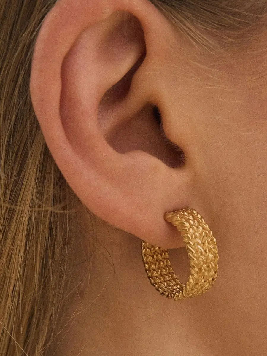 Agapée Kotsi wide gold hoop earrings featuring a textured, asymmetrical braid design worn on an ear.