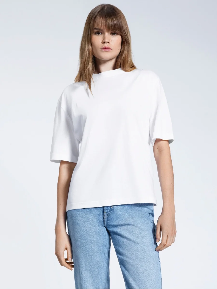 A woman wears a thick, structured white crew-neck organic cotton t-shirt from Plain and Simple tucked into light-wash denim jeans.