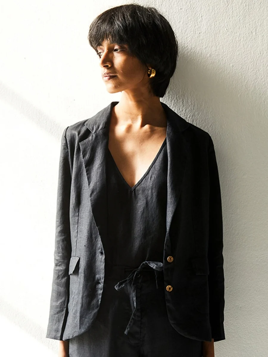An elegant, front-facing medium shot of a woman with short, dark hair looking off to the left against a white wall. She is wearing a structured, charcoal black linen blazer with notched lapels and brown buttons over a matching V-neck linen camisole and drawstring trousers. Chunky gold hoop earrings catch the light, adding a polished accent to her minimalist, sophisticated outfit.