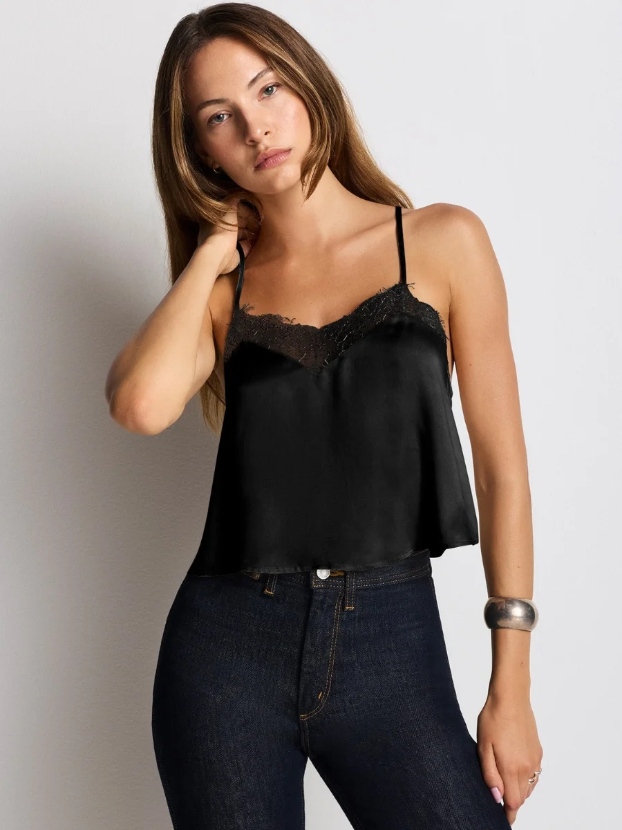 A woman modeling a black bias-cut silk Tencel blend camisole from RE/DONE featuring delicate lace trim along a straight neckline, paired with high-waisted dark wash denim jeans.