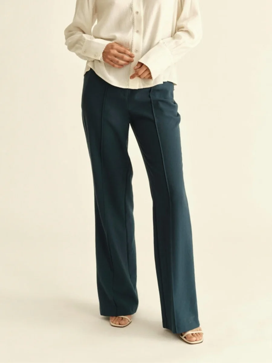 A woman modeling a cream silk long-sleeve blouse tucked into high-waisted deep teal straight-leg tailored trousers from Cartolina with front pintuck details.