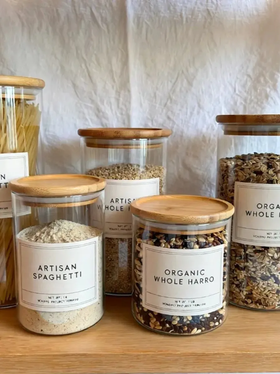 Clear glass food storage canisters with light brown wooden lids and clean square labels filled with dry grains and pasta sitting on a shelf against a white textured fabric background.