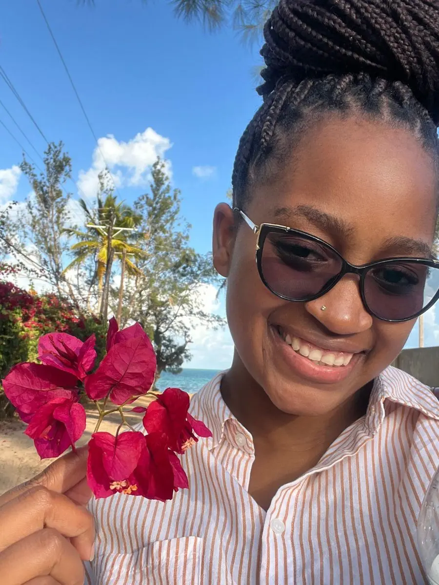 A smiling woman wearing black cat-eye sunglasses and a striped button-down shirt, holding bright pink bougainvillea flowers with her hair styled in a large braided high bun.