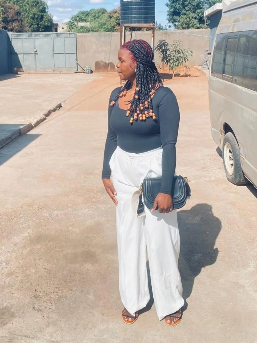 A woman standing outdoors in a black long-sleeve top and high-waisted white wide-leg trousers, showcasing mid-length box braids with chunky wooden beads on the ends.