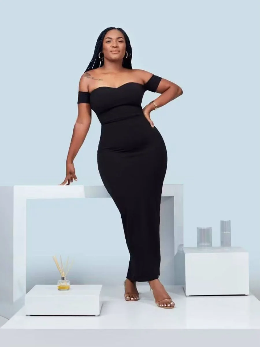 • A woman standing in a studio setting wearing a fitted, floor-length black off-the-shoulder gown, showcasing long knotless box braids with a clean center parting falling over her shoulders.