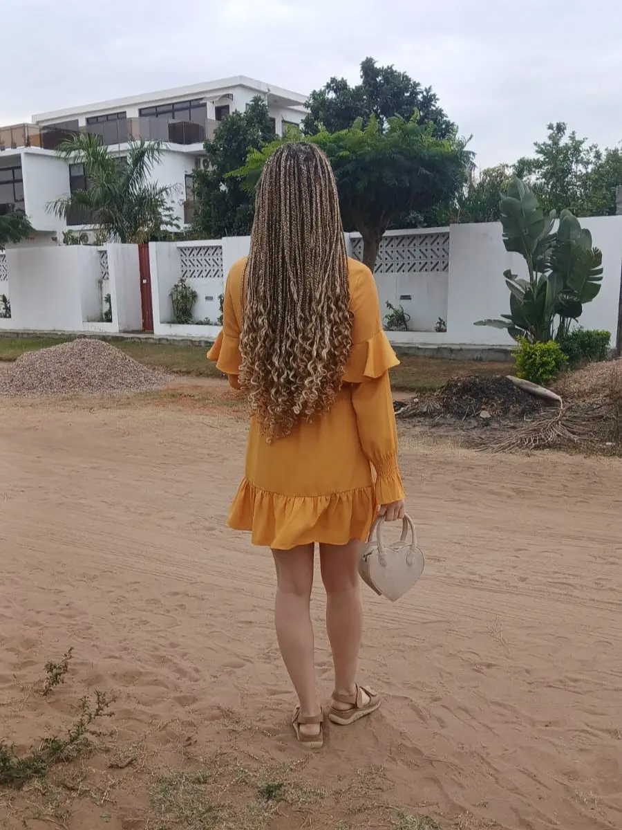 A view from behind of a woman walking outdoors in a long mustard yellow ruffle minidress and sandals, holding a white heart-shaped bag, showcasing long knotless braids with curly blonde-highlighted ends cascading down her back.