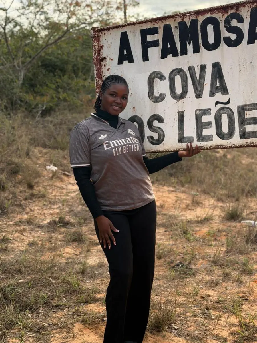 • A woman standing outdoors in front of a rustic sign, wearing a grey sports jersey layered over a black turtleneck sweater and black pants, showcasing tight cornrow braids braided flat against her scalp.