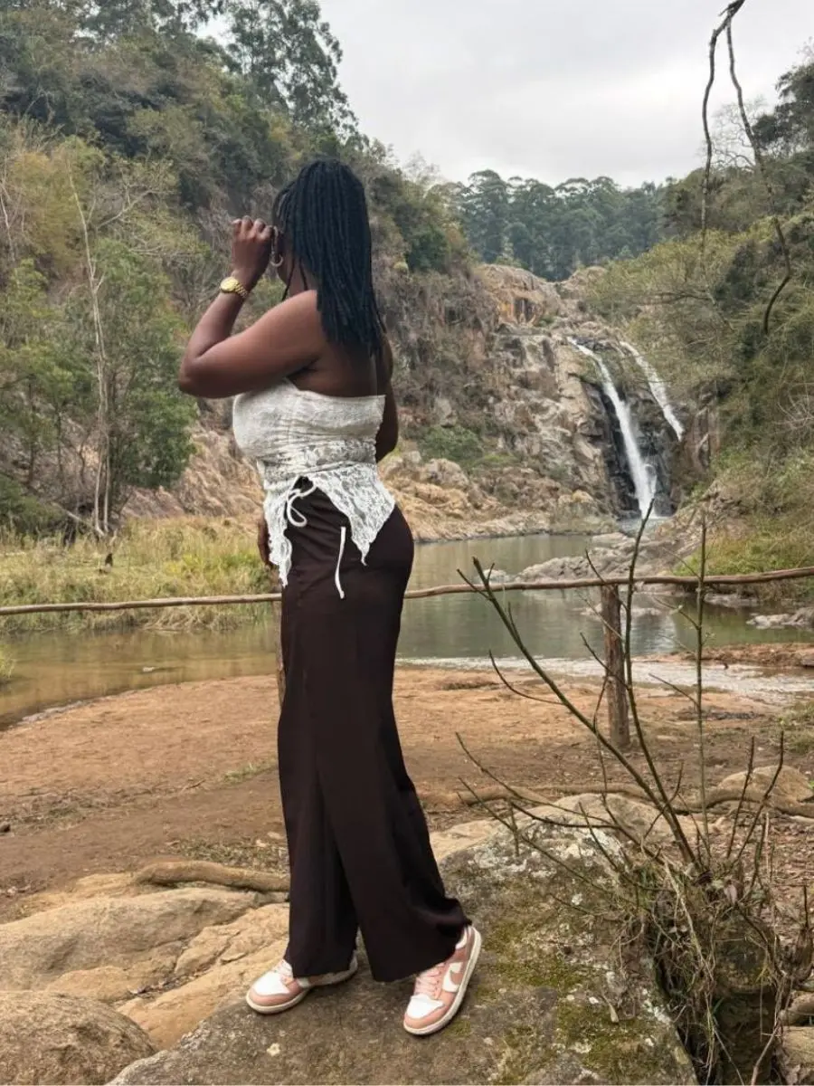 A woman standing on a rock by a waterfall wearing a white lace strapless tube top, dark fluid wide-leg trousers, and sneakers, showcasing shoulder-length twist braids tied in a half-up style.