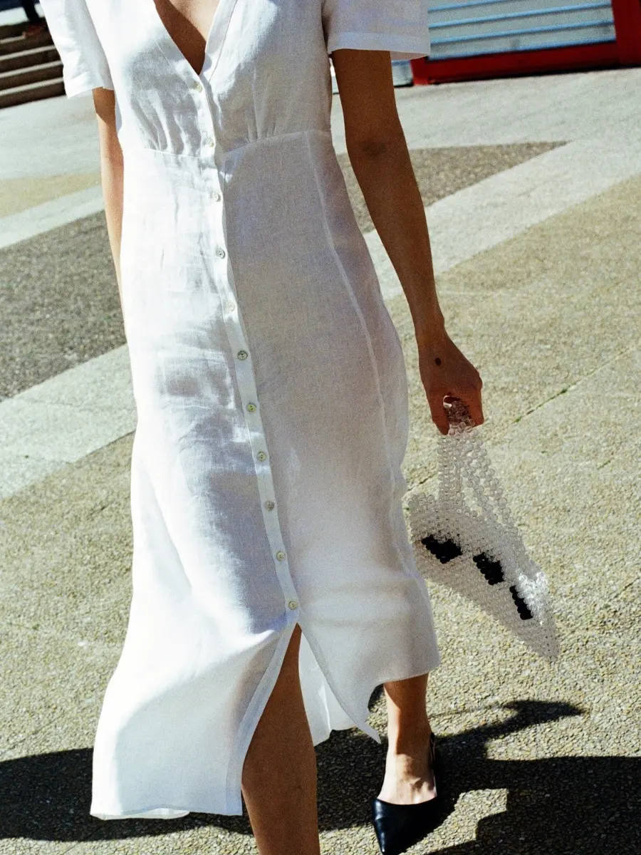 A woman walking outdoors wearing a white midi-length linen shirt dress from Má + Lin featuring a button-down front, styled with black pointed-toe slingback flats.