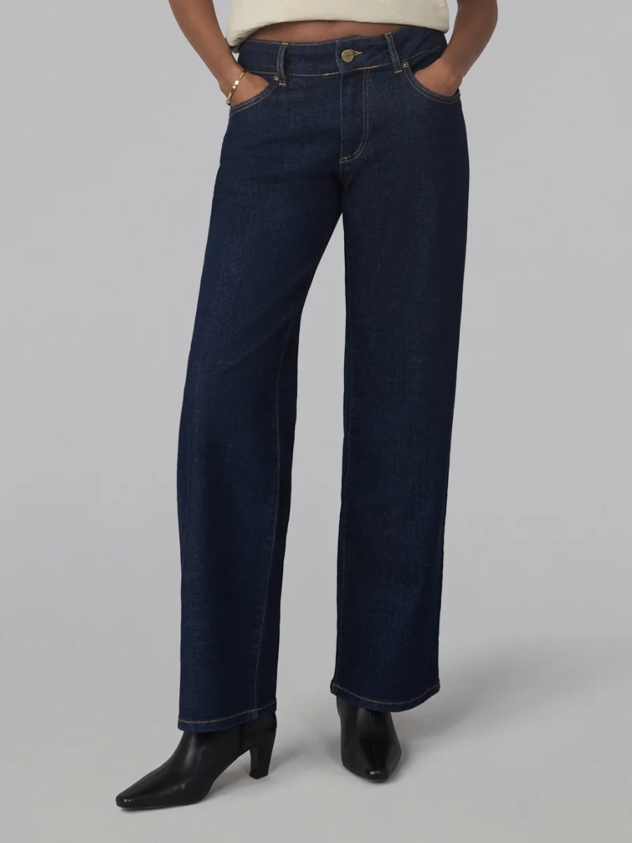 A model standing against a grey background, showcasing a pair of dark rinse indigo straight leg jeans from Lola Denim styled with black leather heeled ankle boots.