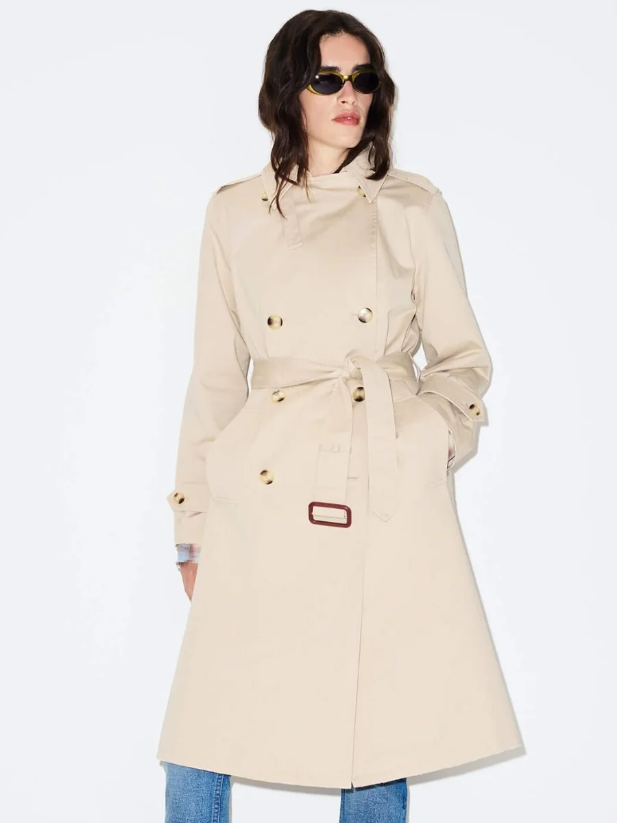 A model posing against a clean white background, showcasing a double-breasted beige cotton twill trench coat from RE/DONE cinched at the waist with a self-belt and styled over blue jeans.